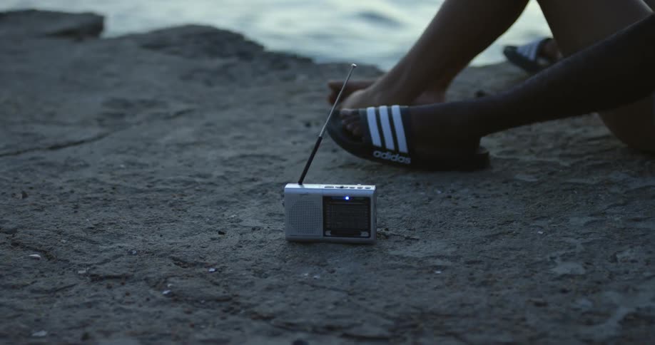 A couple is sitting on concrete on the waterfront listening to a radio