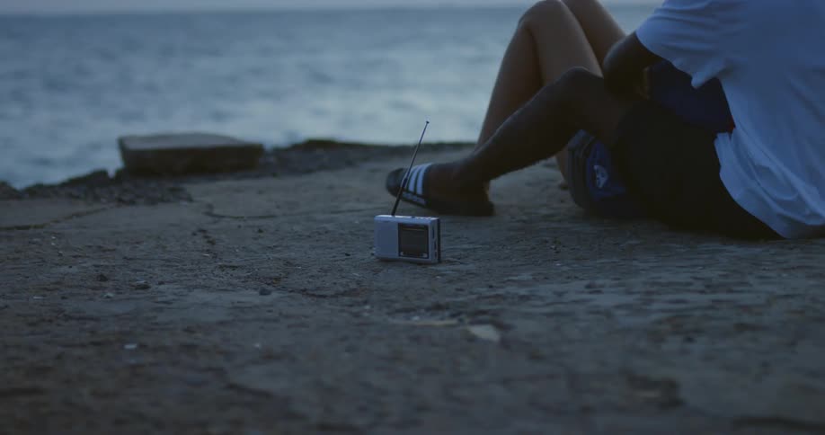 Man with white t shirt black shorts and slippers holds woman with shorts sitting on concrete pier and listening to radio