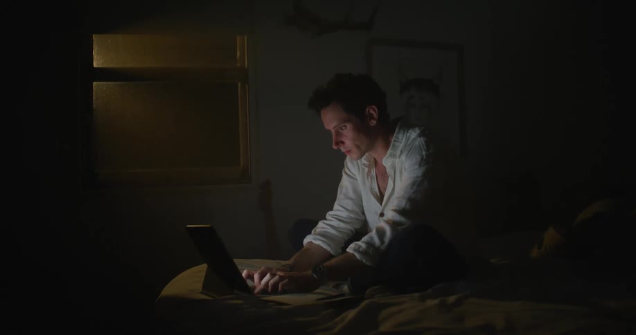 A man sits on his bed and types on a laptop and then covers his face with his hands and rubs his eyes