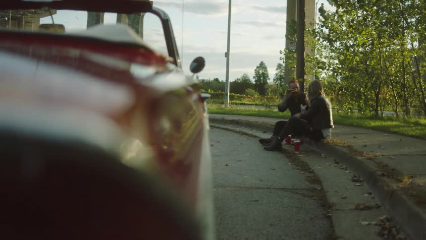 Man and woman sit on curb eating near parked convertible as another car passes in background