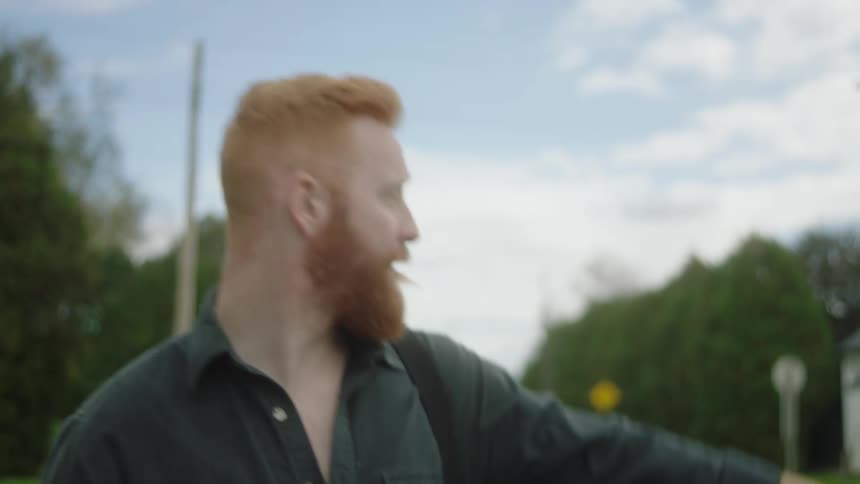 Red haired bearded musician carries instrument case while hitchhiking along road through neighborhood