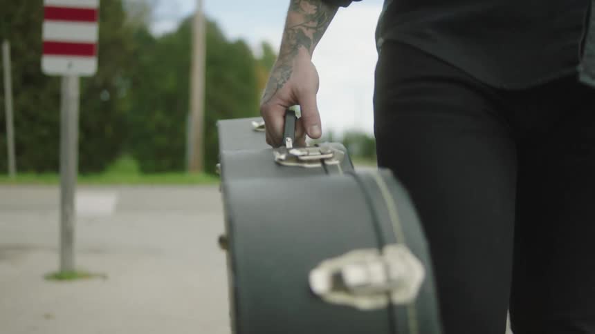 Male musician carries instrument case along sidewalk near intersection