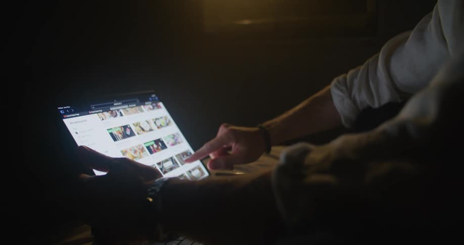 A man is sitting and scrolling on a laptop screen in a dark room at night