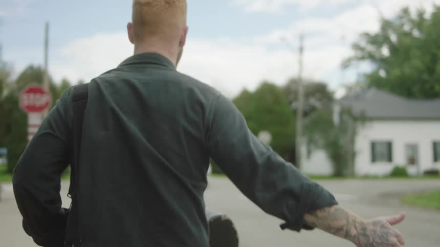 Young blonde bearded man carrying a guitar case hitchhiking on a small town road