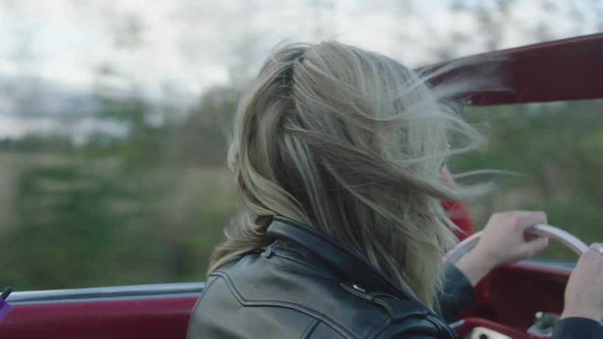 A woman is driving a red vintage convertible car on a cloudy day
