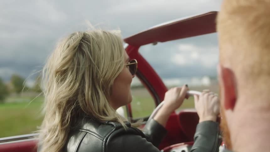 Blonde woman wearing sunglasses and black leather jacket driving a red vintage car on the countryside