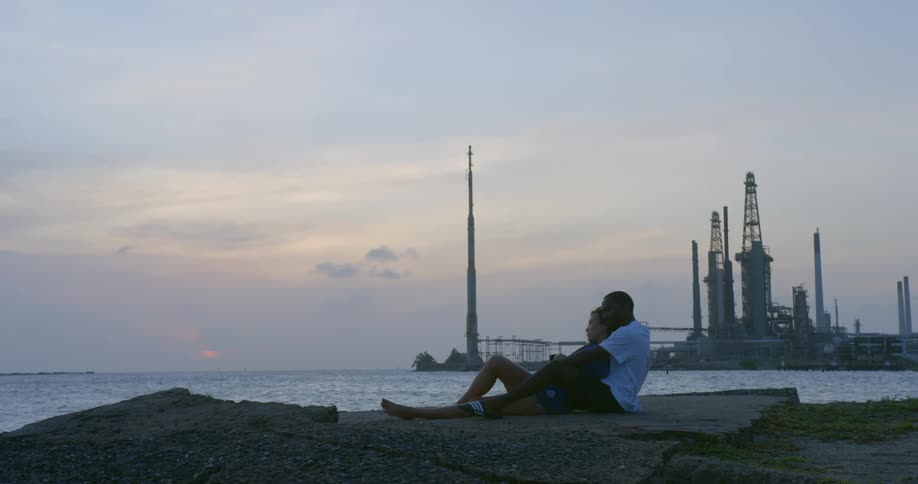 Male back hugs woman as they sit on shore with factory in background