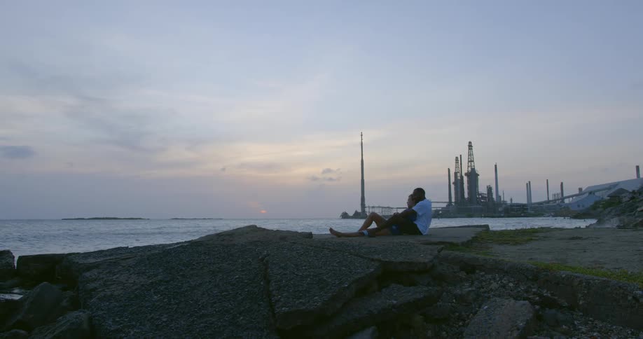 Two people are sitting on broken concrete on a coast looking out at the water