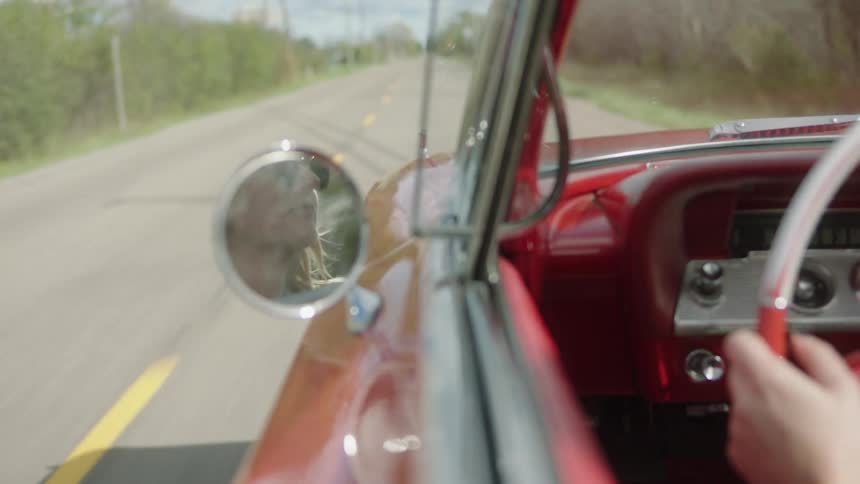 Side mirror of vintage car reflect blonde female driver as car moves through countryside