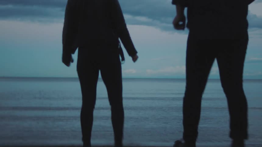 Man and woman run across beach to surf then stand smiling and talking as moon peaks through clouds