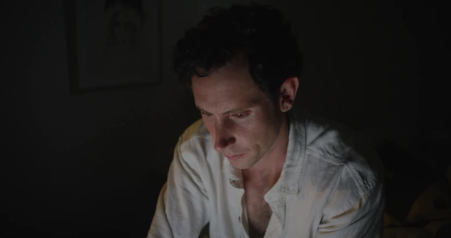 A mans face is lit up in a dark room by his laptop as he types while sitting in bed
