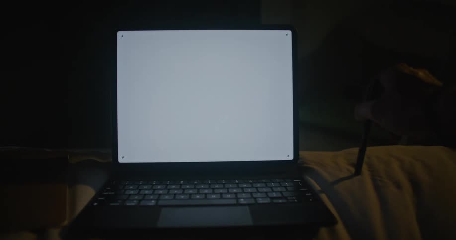 A person is sitting in front of a blank laptop screen at night and fidgeting with a pen