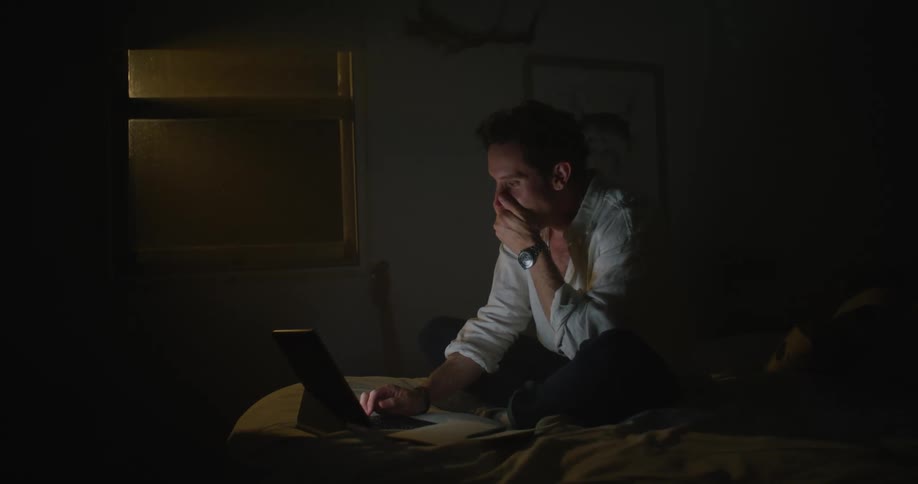 Man sits cross legged on bed in dark room yawning as he types on laptop