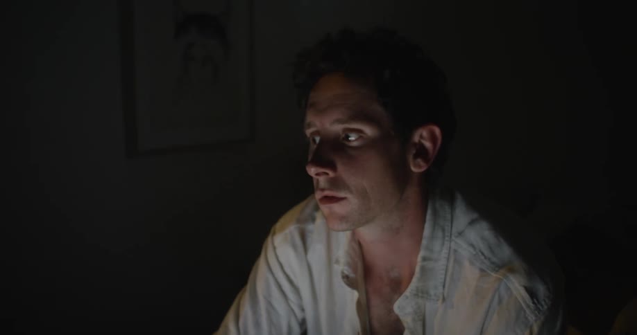 Man sits cross legged on bed in dark bedroom typing on laptop