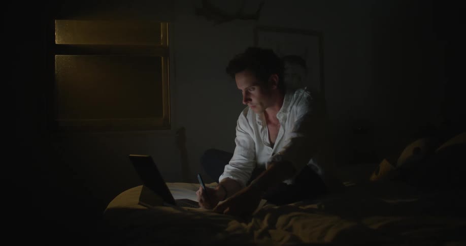 A man is sitting on a bed in a dark room typing on a laptop and writing on a notepad