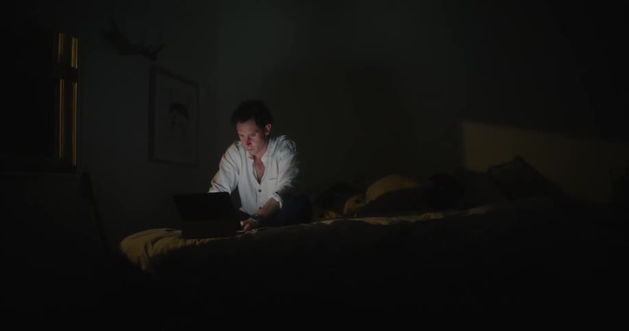 Man with white shirt sits on bed and uses laptop in dark bedroom
