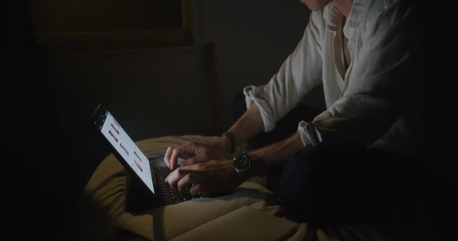 A man is sitting on a bed thinking and typing on a laptop in a dark room