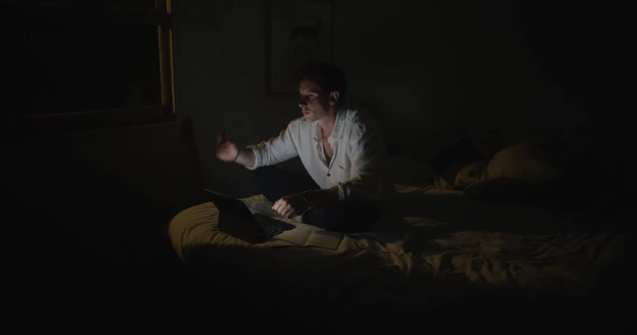 A man sitting cross legged on top of his bed working on a laptop