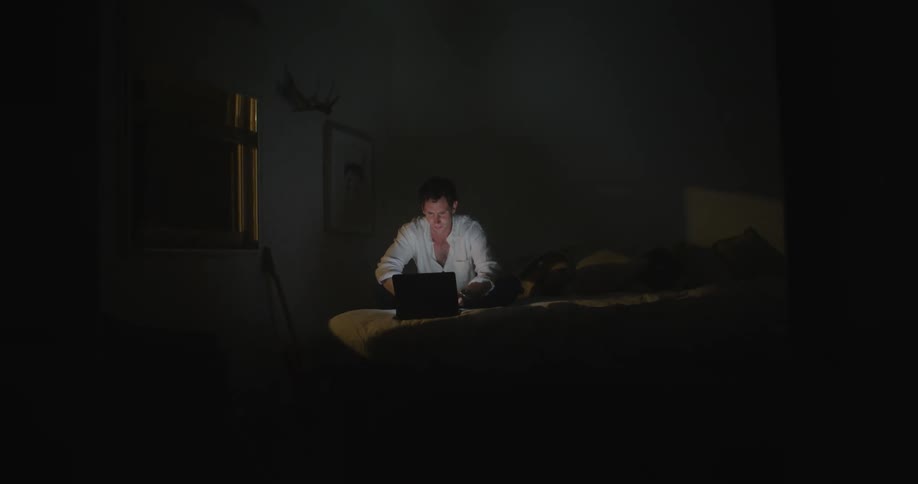 A man closes a laptop he is using and lays down in bed in a dark bedroom