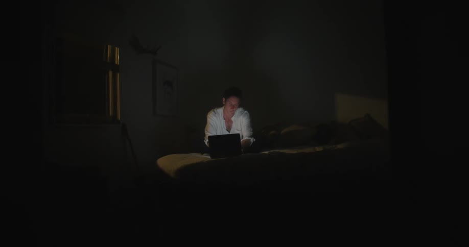 A man is working late on his laptop in his bedroom at night