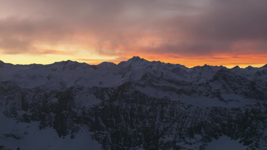 An aerial view of snowy mountains and low clouds at dusk with an orange sky