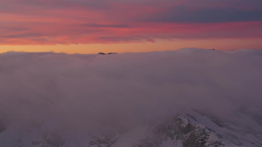 A layer of clouds covering snow covered mountain peaks and ridges with a colorful sunset sky above
