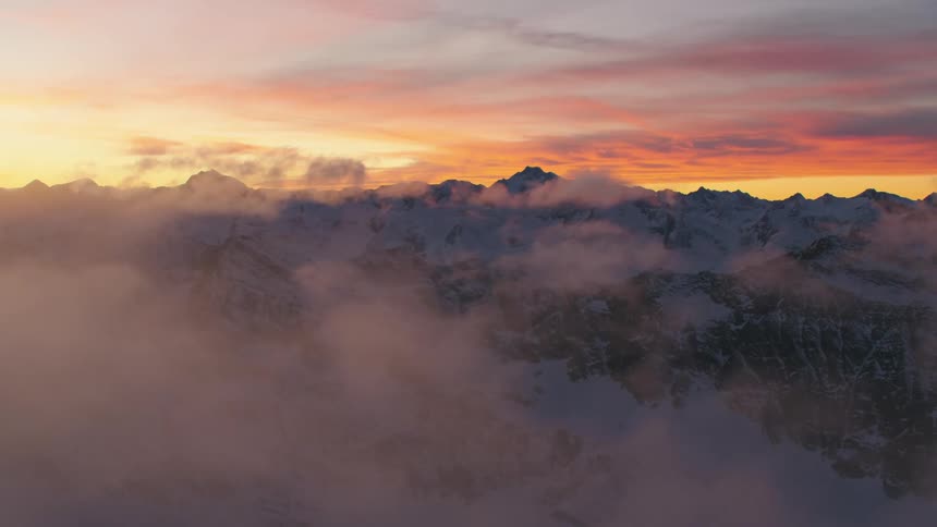 Clouds move by snow covered mountains with peaks high in a red cloudy sky