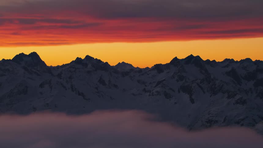An aerial view of snowy mountains with low clouds and an orange sky during a sunset
