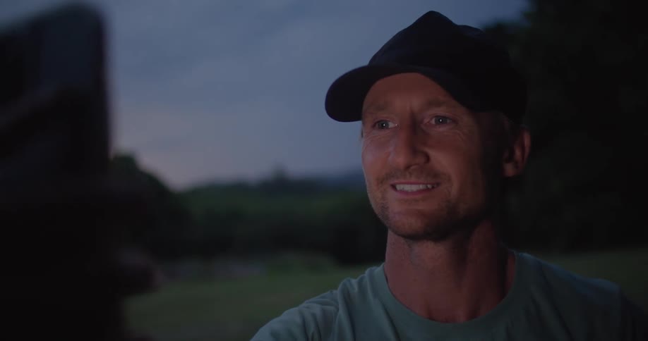 A man holding up his phone waves smiles and talks during a video call at sunset