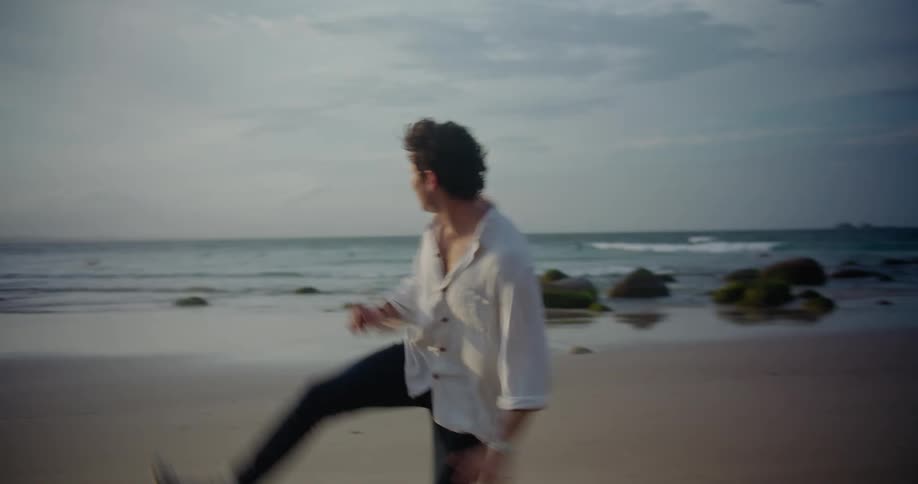 A man is walking on a beach and picks up a rock and throws it into the ocean