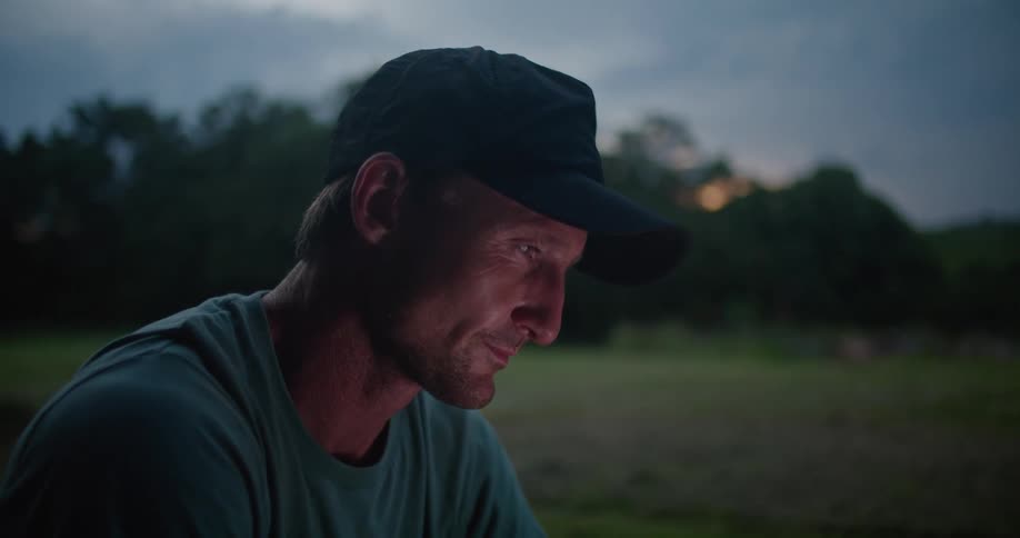 A man is sitting in a grass field at dusk while using a smartphone