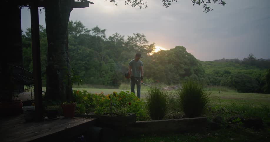 A man moves a gas powered trimmer back and forth near a house deck and planters