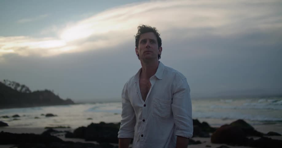 A man starts to stretch then puts his hands on his face as he stands on a rocky beach