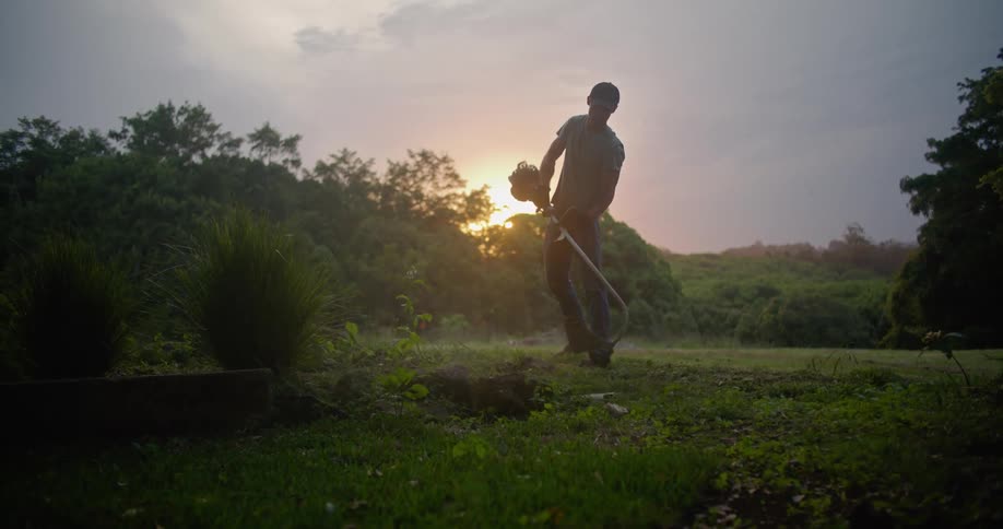 Man cuts overgrowth using gas powered weed trimmer silhouetted by back light from sun on horizon in red sky
