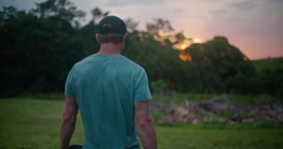 A man is walking away carrying a shovel and a bucket as the sun is setting behind the trees