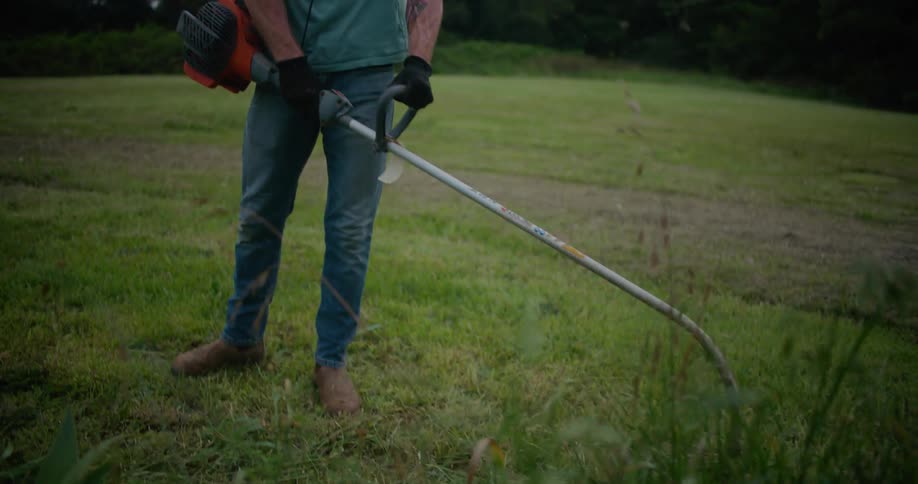 A man uses a weed wacker to trim grass around a bush