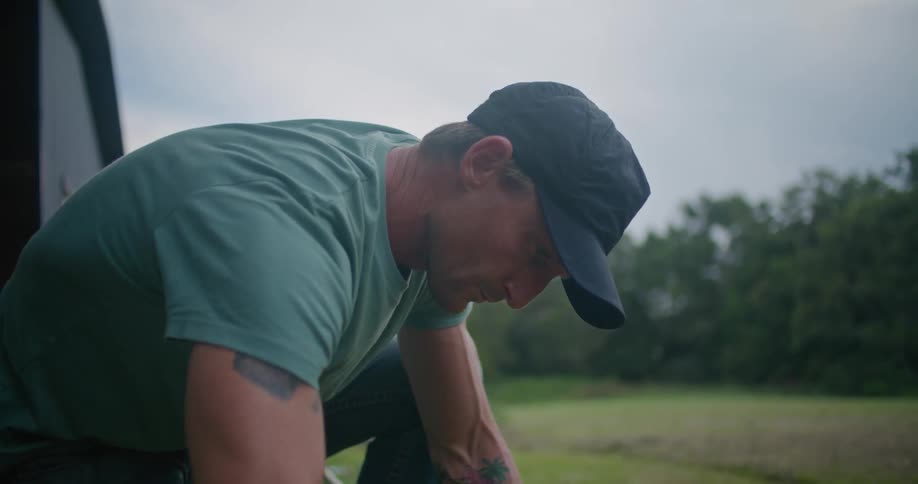 A man wearing a black baseball cap and gloves is kneeling down on the grass and fiddling with an electric weed trimmer