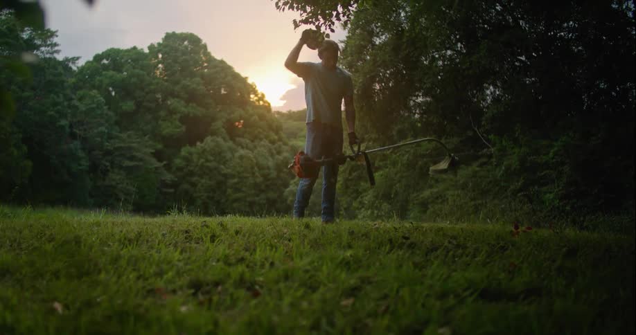A man is using a string trimmer then in a field then removes his hat and kneels in the grass