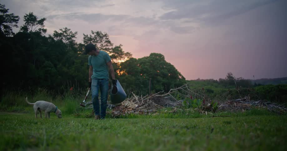 A man is walking in a field with a dog and carrying a shovel and bucket