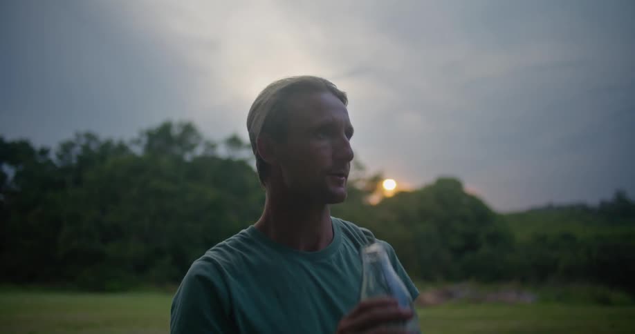A man standing in a grassy field takes a drink of water and then takes his hat off and wipes his forehead