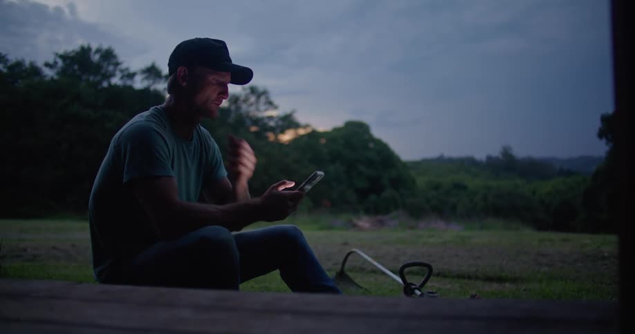 A man is sitting on the grass looking at his phone and he takes a drink from a bottle