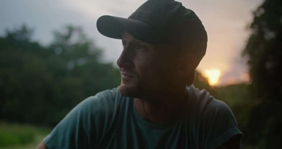 A man wearing a baseball cap is looking around and takes off his cap as the sun is setting behind the trees