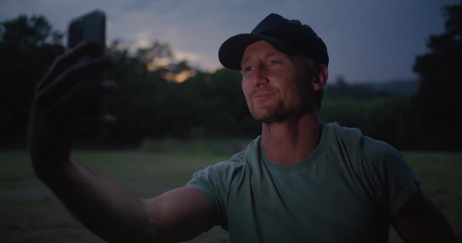A man is video chatting on his phone while sitting in a grass field at dusk