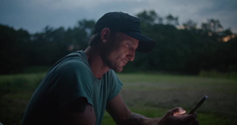 A man is sitting in a field using a smartphone with lawn care equipment nearby late in the evening
