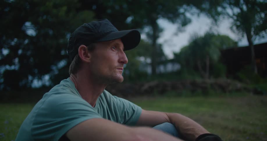 A man wearing gardening gloves is sitting on a grassy lawn and staring off