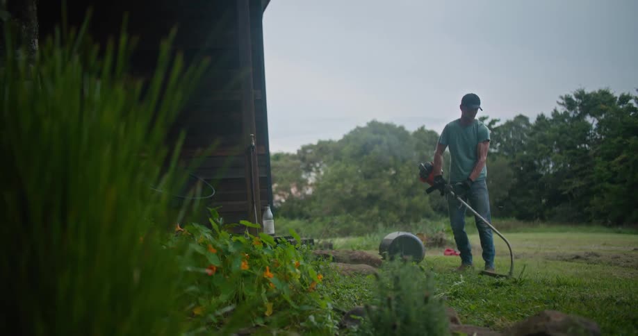 A mans arm muscles strain as he uses a gas powered trimmer to cut back weeds and grass near an out building