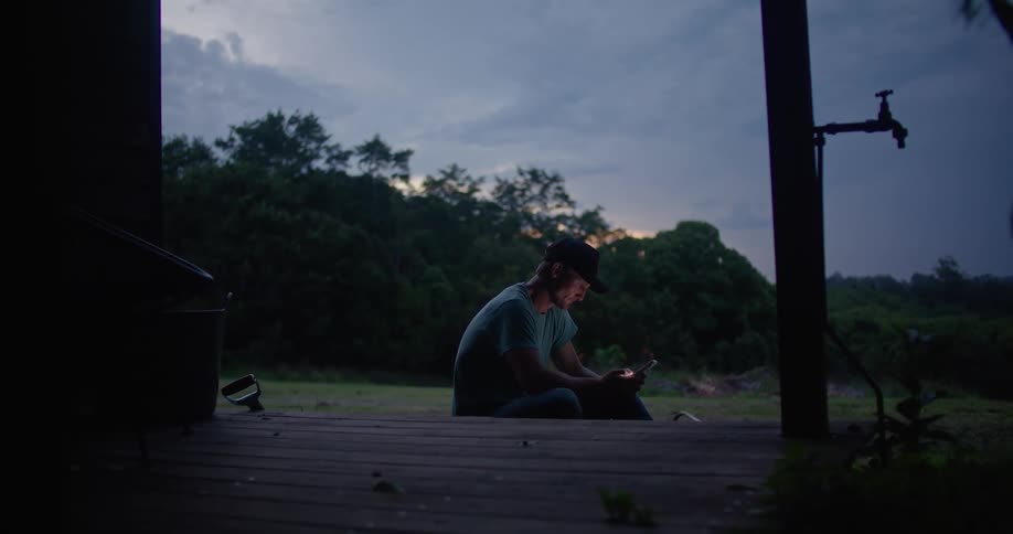 A man sitting outside near a porch deck scrolling on his phone and looking up at the sky