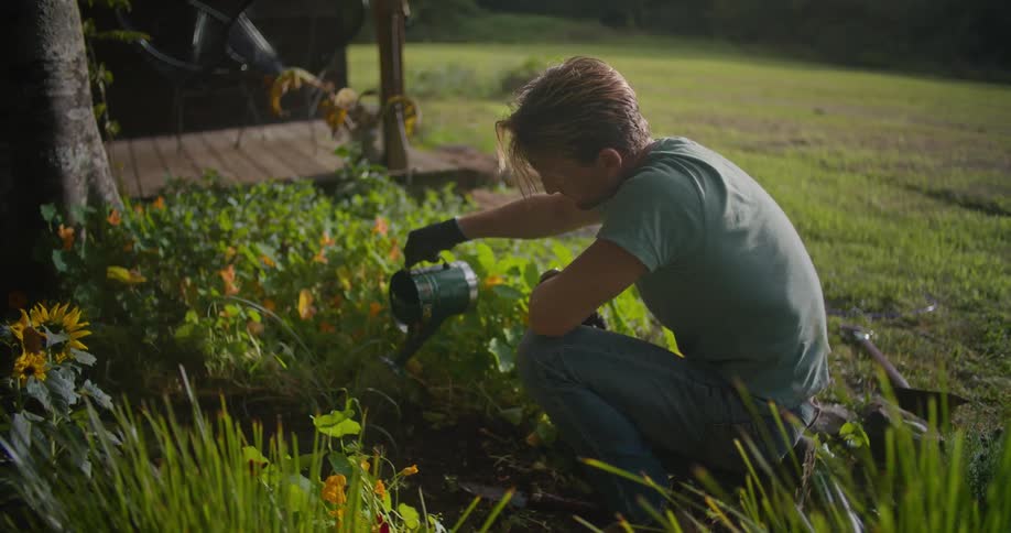 A man kneels on the ground and waters flowers next to his porch