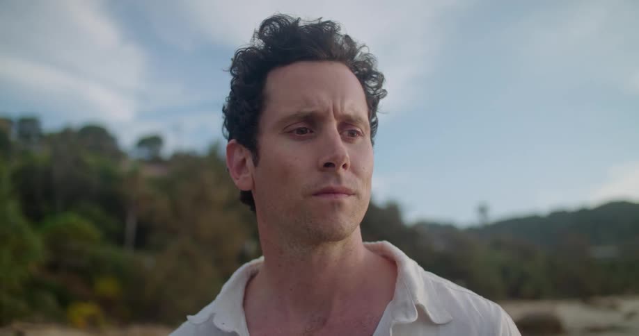 Man wearing white button up shirt stands on beach on windy day with mountains and woods in background