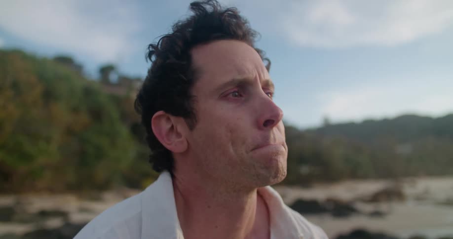 A man sitting outside on the sand sniffles and sighs as he tries to keep himself from crying
